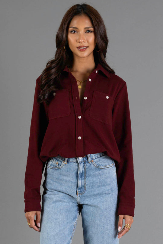 A woman with long wavy hair wears the Panache Apparel Collection Burgundy Soft Button Up, featuring chest pockets, paired with light blue jeans. She stands against a plain gray background, smiling softly at the camera.