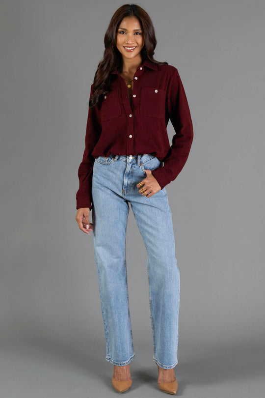 A woman stands against a gray background wearing the Panache Apparel Collection Burgundy Soft Button Up, paired with light blue jeans and tan heels; her long wavy hair frames a bright smile.