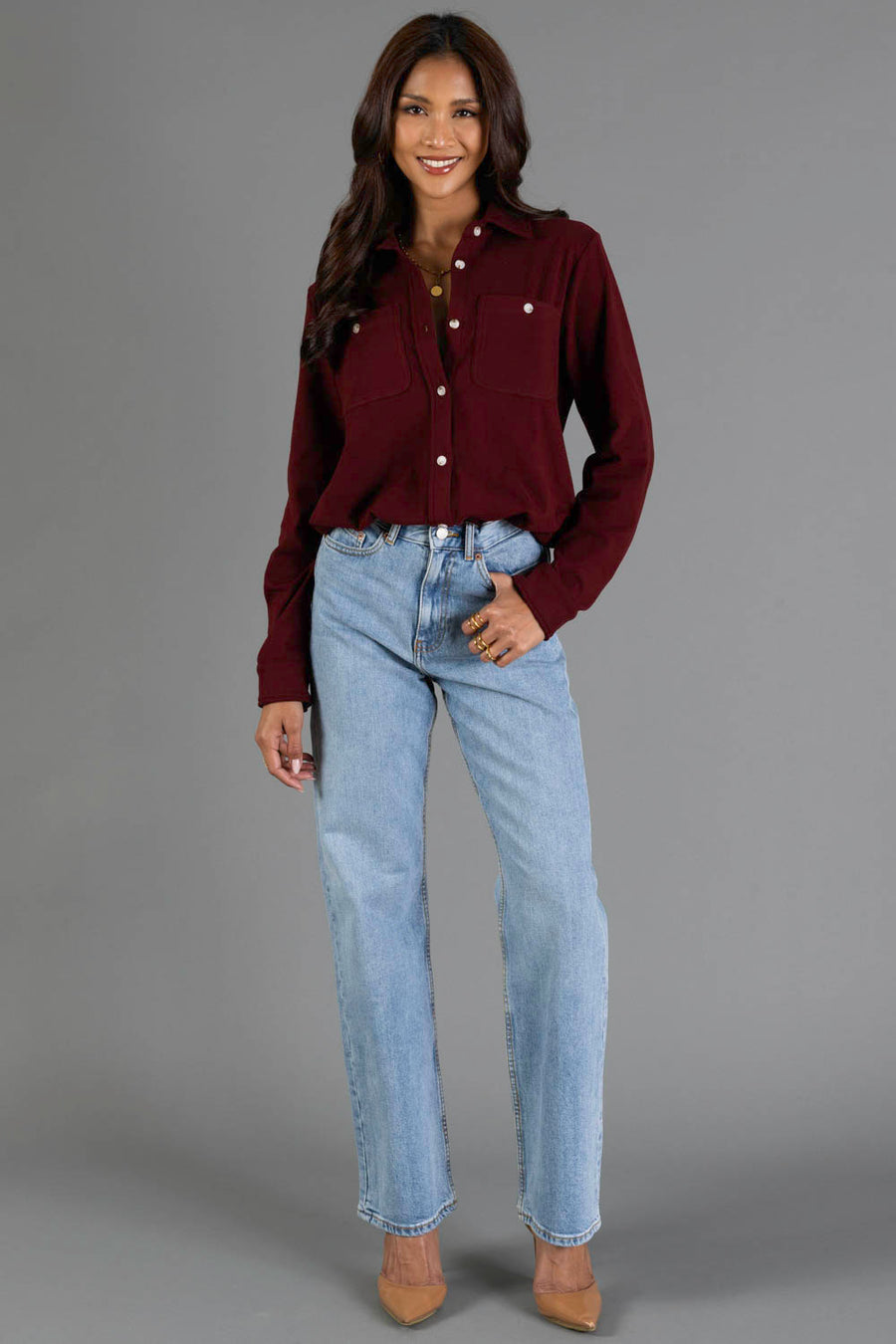A woman stands against a gray background wearing the Panache Apparel Collection Burgundy Soft Button Up, paired with light blue jeans and tan heels; her long wavy hair frames a bright smile.