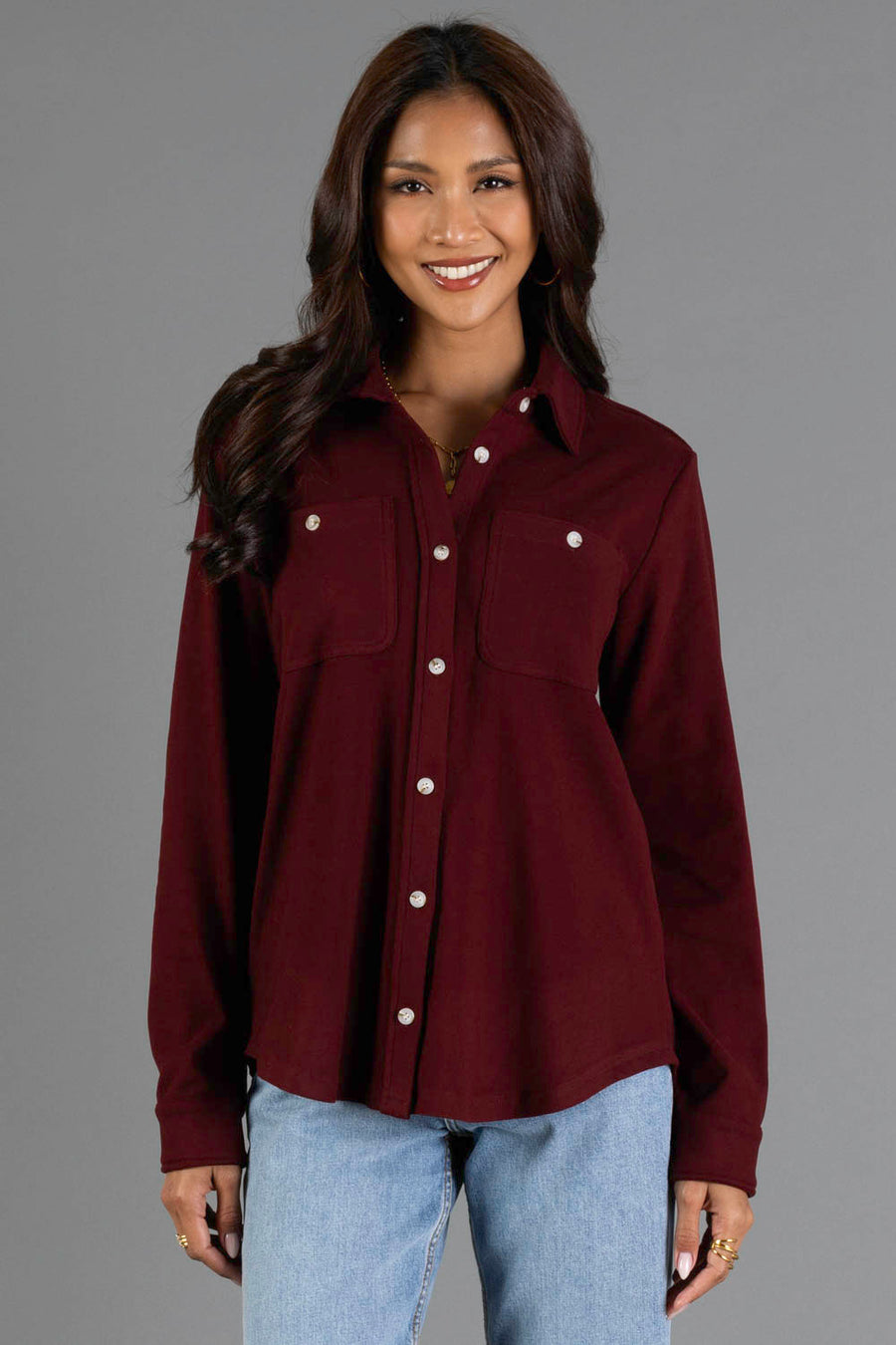 A woman stands and smiles in front of a plain gray background, wearing the Panache Apparel Collection Burgundy Soft Button Up with chest pockets, paired with light blue jeans.