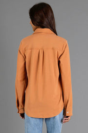 A woman with long dark hair, wearing the Muted Orange Soft Button Up from Panache Apparel Collection and light blue jeans, stands facing away from the camera against a plain gray background.