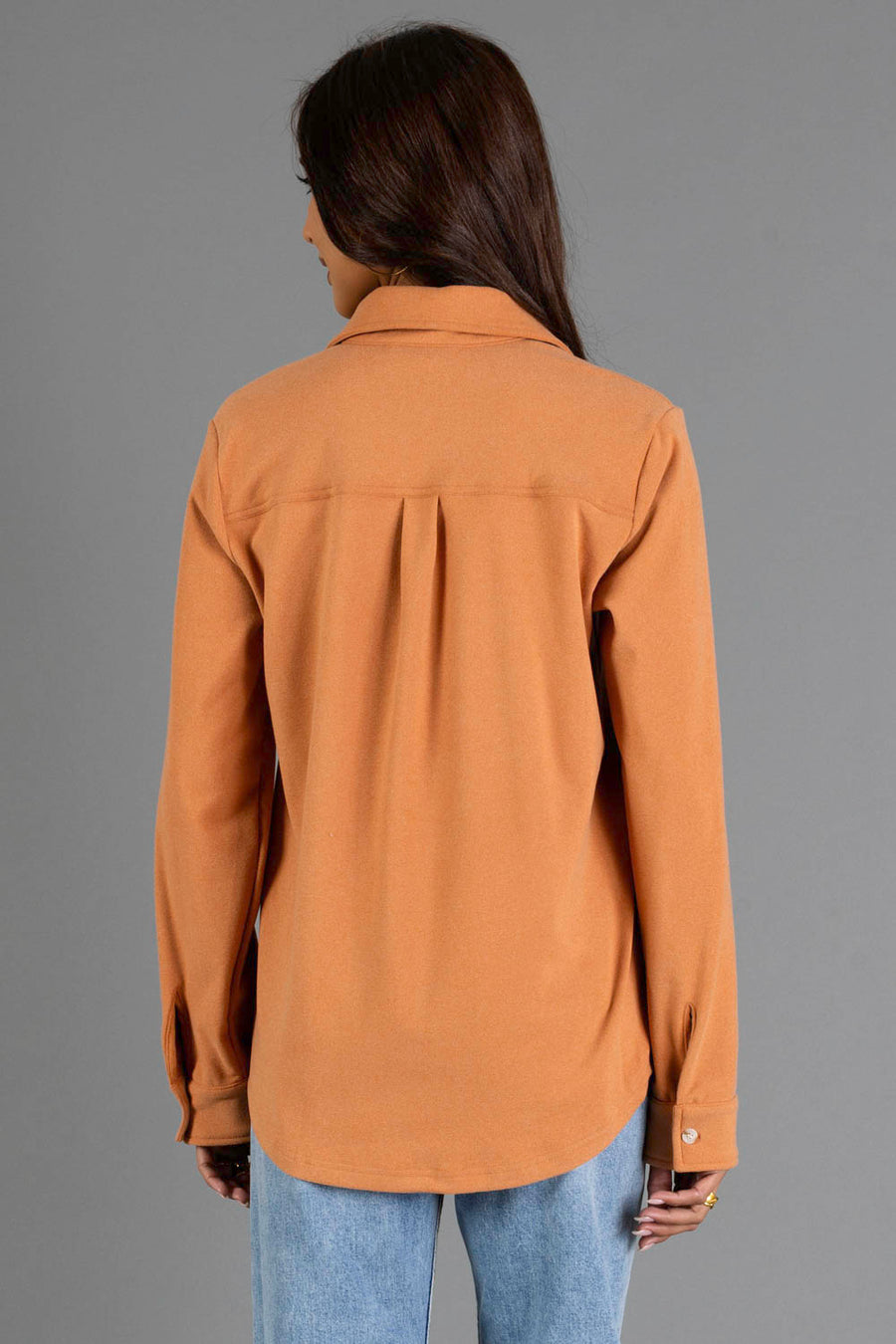 A woman with long dark hair, wearing the Muted Orange Soft Button Up from Panache Apparel Collection and light blue jeans, stands facing away from the camera against a plain gray background.