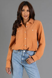 A woman wearing the Panache Apparel Collection Muted Orange Soft Button Up with long sleeves and front pockets, paired with light blue jeans, stands against a plain gray background with one hand in her pocket.