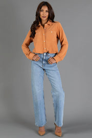 A woman stands against a gray backdrop wearing the Muted Orange Soft Button Up from Panache Apparel Collection, paired with light blue high-waisted jeans and beige heels. She has long, wavy hair and her hands rest on her hips.