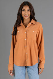A woman with long, wavy brown hair wears the Panache Apparel Collection Muted Orange Soft Button Up, featuring chest pockets, paired with light blue jeans as she stands and smiles against a plain gray background.