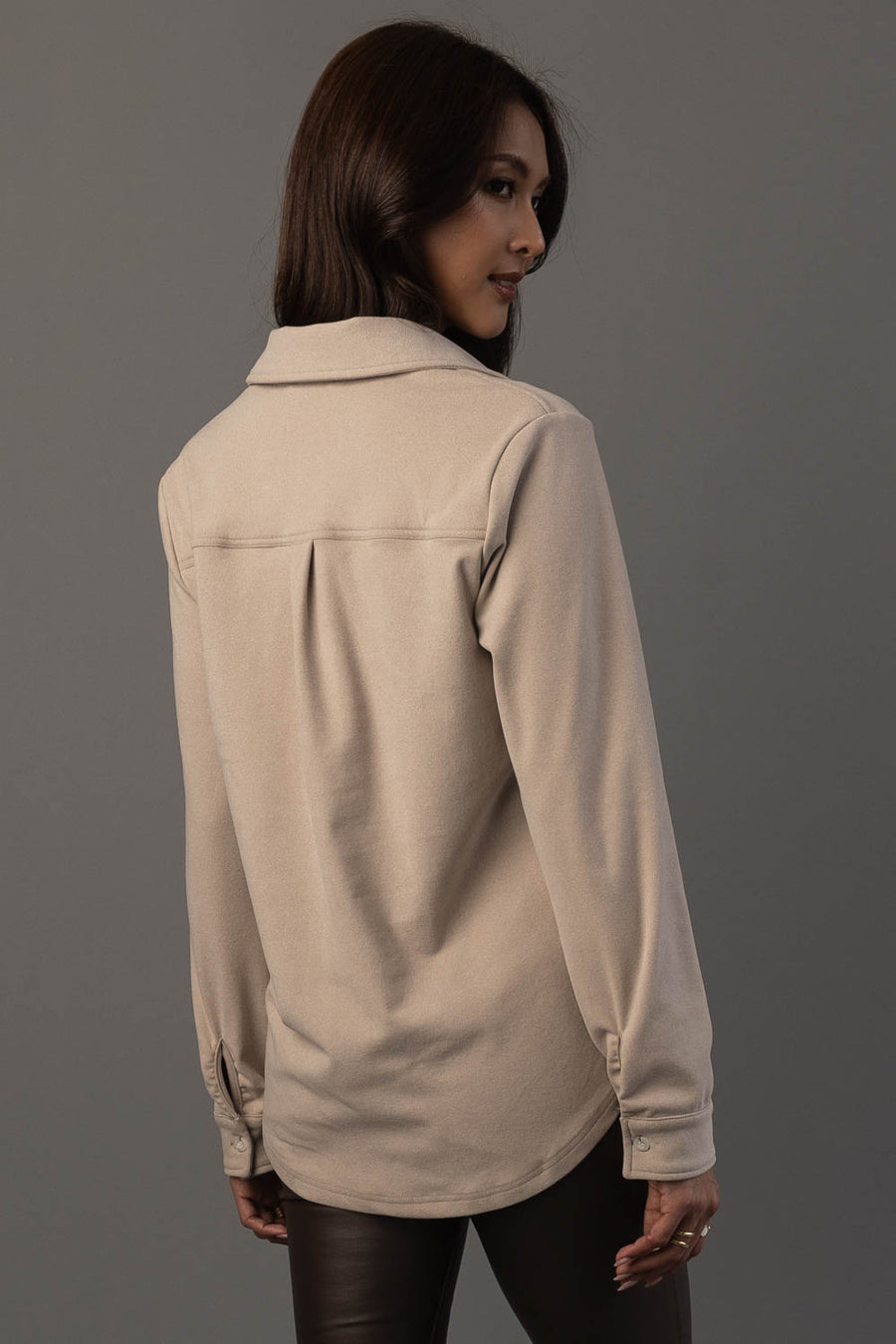 A woman with long dark hair is standing in front of a gray background, facing away and looking over her shoulder. She is wearing the Light Tan Soft Button Up from Panache Apparel Collection and dark pants.