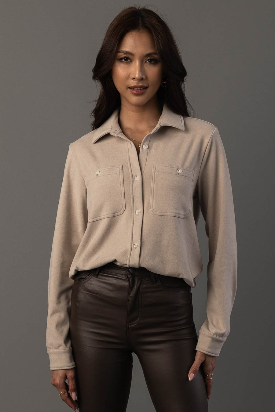 A woman with long dark hair wears the Panache Apparel Collection’s Light Tan Soft Button Up, a beige shirt featuring two front pockets, paired with dark brown leather pants, standing against a plain gray background.