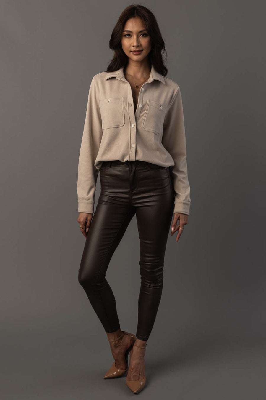 A woman stands against a gray background, wearing the Light Tan Soft Button Up from Panache Apparel Collection, paired with black leather pants and tan pointed-toe heels. She has long dark hair and a relaxed, confident pose with one leg slightly bent.