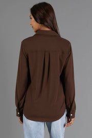 A woman with long dark hair, wearing the Panache Apparel Collection Brown Soft Button Up and light blue jeans, is standing with her back to the camera against a plain gray background.
