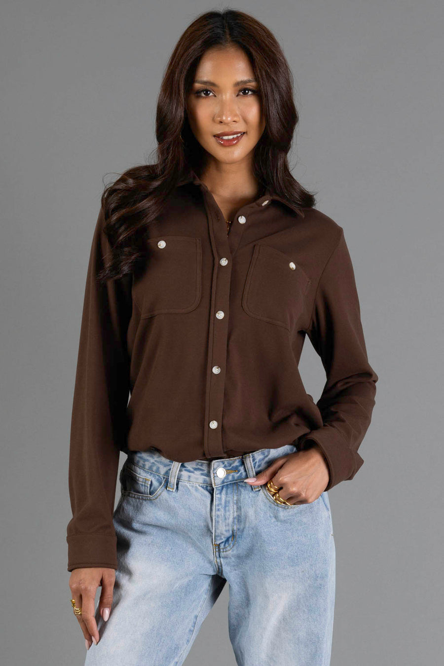 A woman with long dark hair is seen standing against a plain gray background, smiling with one hand in her pocket. She wears the Panache Apparel Collection Brown Soft Button Up, featuring front pockets, paired with light blue jeans.