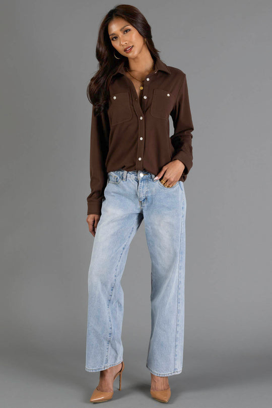 A woman with long dark hair poses against a gray background with one hand in her pocket, wearing the Brown Soft Button Up from Panache Apparel Collection, paired with light blue high-waisted jeans and beige high heels.
