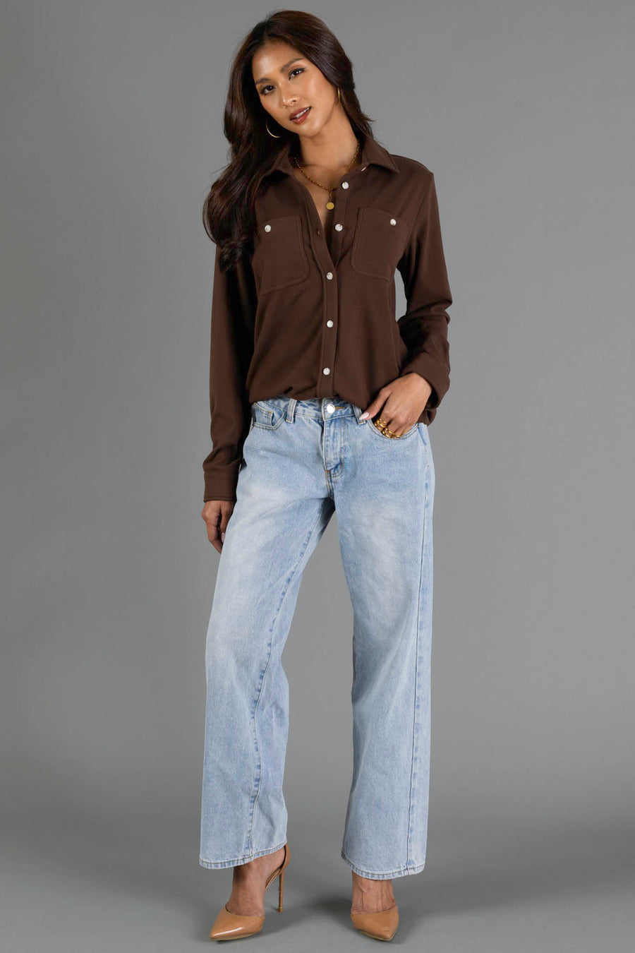 A woman with long dark hair poses against a gray background with one hand in her pocket, wearing the Brown Soft Button Up from Panache Apparel Collection, paired with light blue high-waisted jeans and beige high heels.