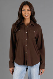 A woman is standing and smiling against a plain gray background wearing the Panache Apparel Collection’s Brown Soft Button Up, a long-sleeved shirt with two chest pockets, paired with light blue jeans.