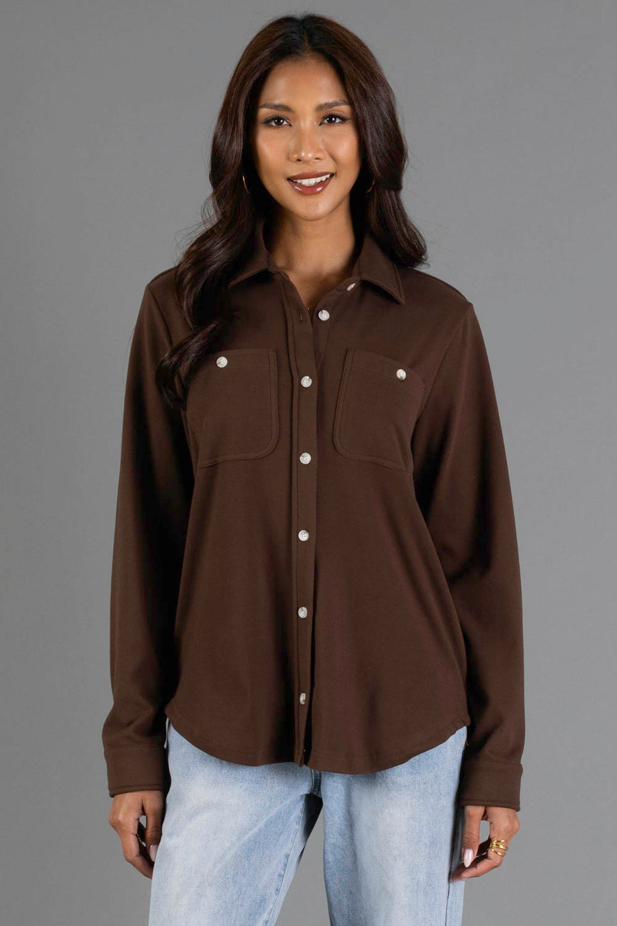 A woman is standing and smiling against a plain gray background wearing the Panache Apparel Collection’s Brown Soft Button Up, a long-sleeved shirt with two chest pockets, paired with light blue jeans.