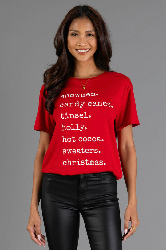 A person wears a red Christmas List Tee by Panache Apparel Collection, a crew neck t-shirt featuring a list of white Christmas-themed words including snowmen, candy canes, tinsel, holly, hot cocoa, sweaters, and christmas.
