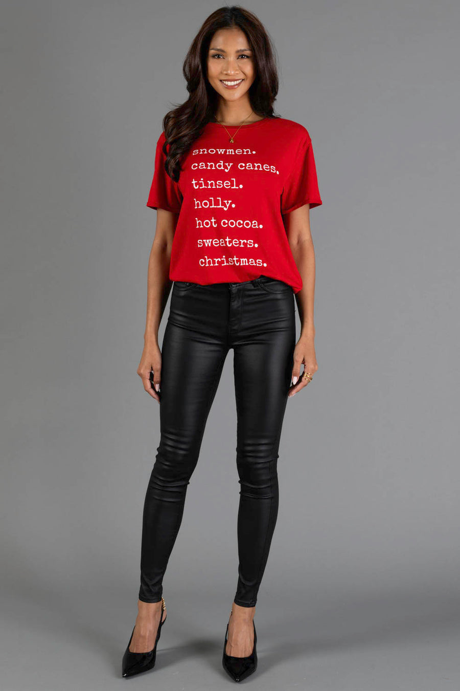 A person wearing a Panache Apparel Collection Christmas List Tee, featuring a red crew neck with white Christmas-themed words, paired with black pants and black heels.