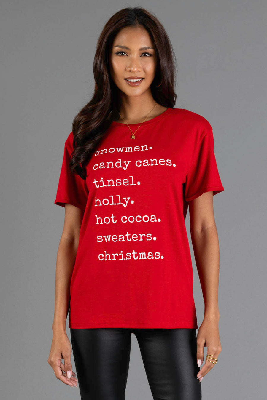 A person wearing a red crew neck Christmas List Tee featuring a graphic print of the words: snowmen, candy canes, tinsel, holly, hot cocoa, sweaters, christmas.