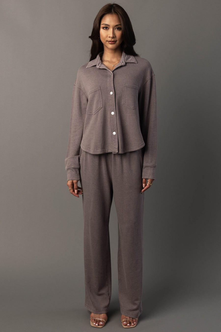 A woman stands against a gray background wearing the Panache Apparel Collection’s Gray Cropped Washed Soft Button Up with matching pants and open-toed heels, looking directly at the camera with a neutral expression.