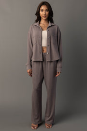 A woman stands against a gray background wearing the Panache Apparel Collection Gray Cropped Washed Soft Button Up, paired with wide-leg pants, a white crop top, and sandals. She has long, dark hair and a neutral expression.