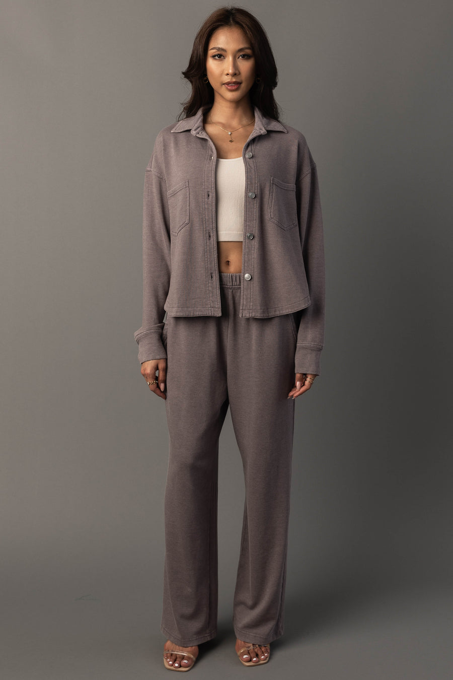 A woman stands against a gray background wearing the Panache Apparel Collection Gray Cropped Washed Soft Button Up, paired with wide-leg pants, a white crop top, and sandals. She has long, dark hair and a neutral expression.