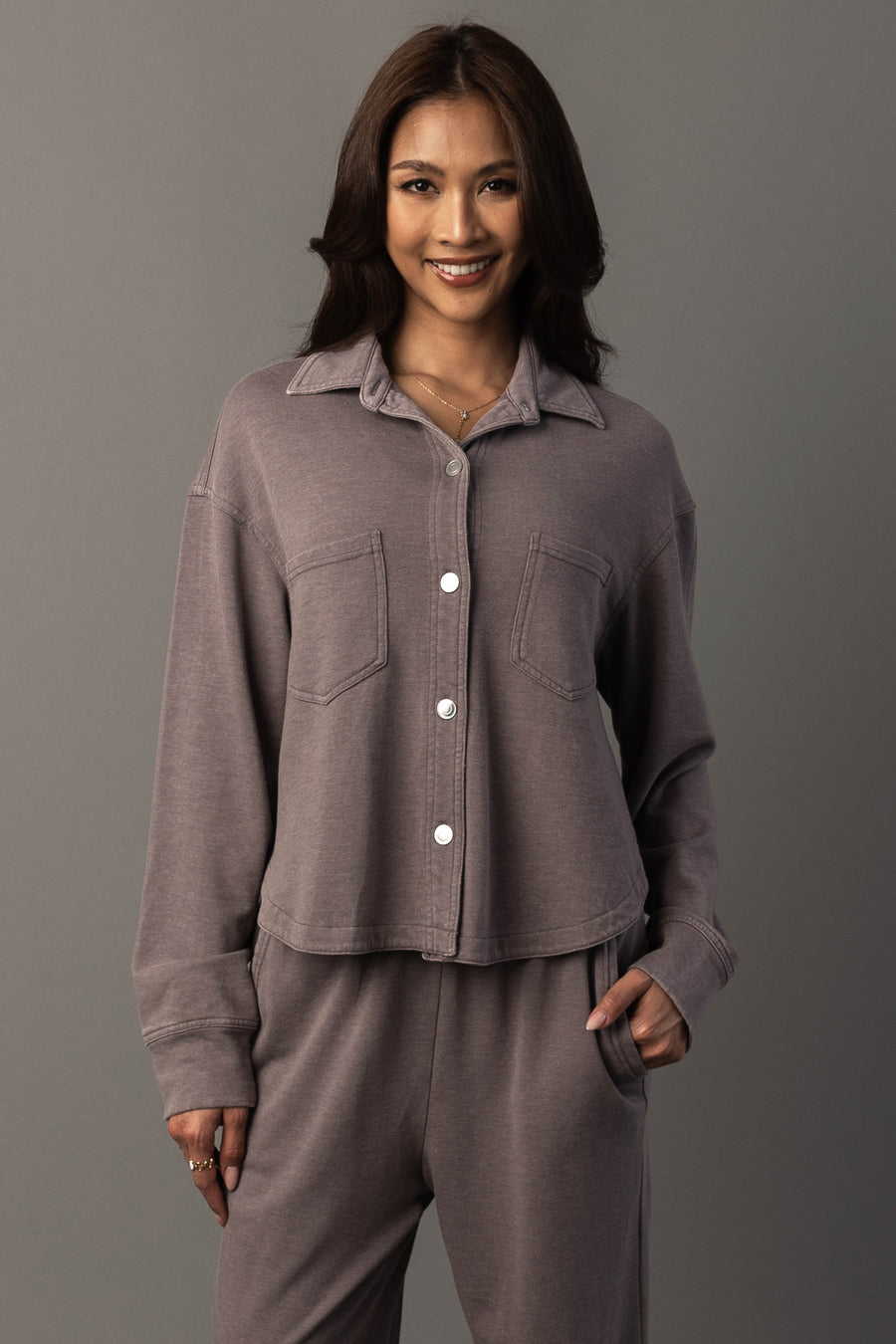 A woman with long dark hair smiles, wearing the Panache Apparel Collection’s Gray Cropped Washed Soft Button Up paired with matching pants. She stands against a plain gray background with one hand in her pocket.