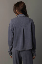 A person with long brown hair, seen from behind, wears the Panache Apparel Collection’s Slate Blue Cropped Washed Soft Button Up and matching pants against a plain gray background.