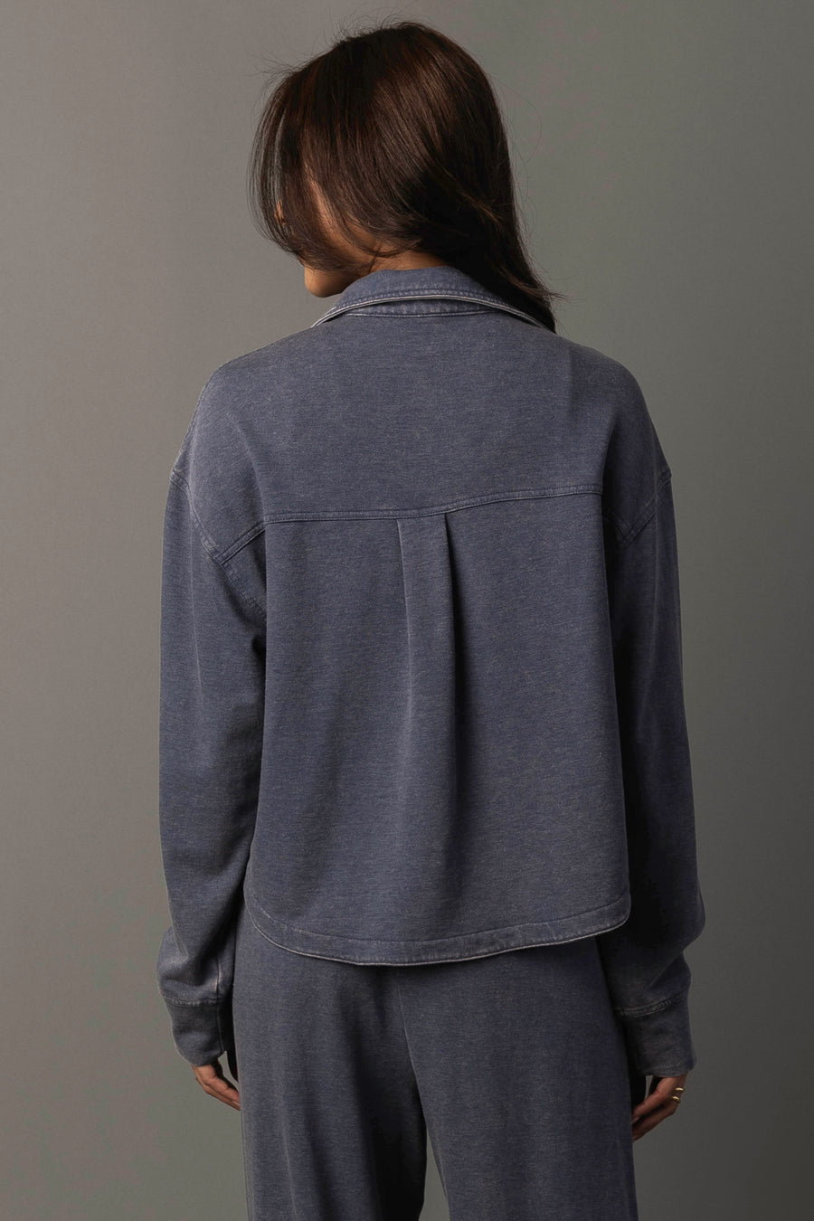 A person with long brown hair, seen from behind, wears the Panache Apparel Collection’s Slate Blue Cropped Washed Soft Button Up and matching pants against a plain gray background.