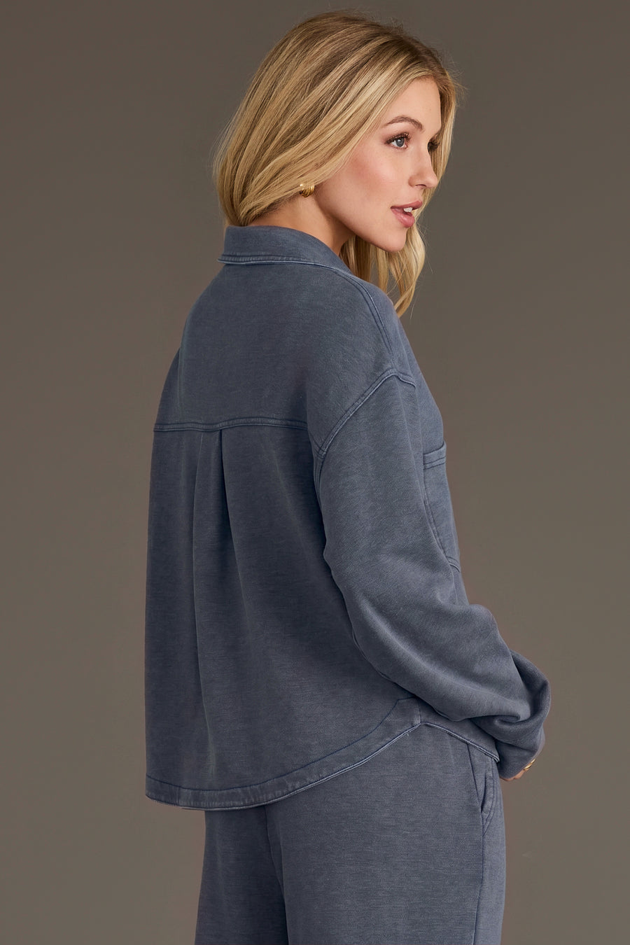 A woman with blonde hair stands sideways against a plain background, thoughtfully gazing to the right, wearing the Panache Apparel Collection Slate Blue Cropped Washed Soft Button Up with matching pants in a relaxed fit.