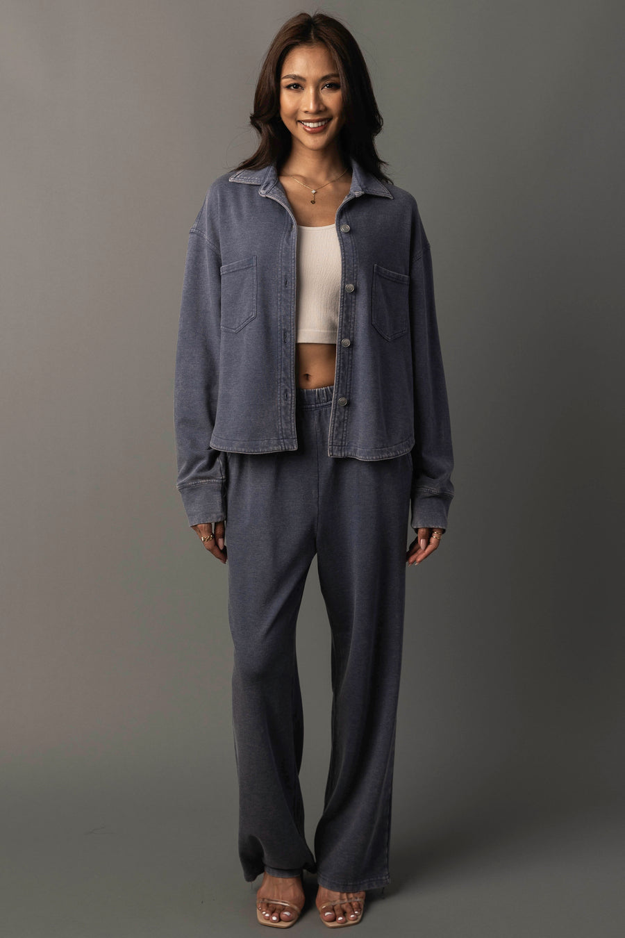A woman stands smiling against a plain grey background, wearing the Slate Blue Cropped Washed Soft Button Up from the Panache Apparel Collection, paired with matching pants over a white cropped top and open-toe heels.