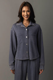 A woman with medium-length dark hair smiles while wearing the Panache Apparel Collection’s Slate Blue Cropped Washed Soft Button Up, paired with matching pants. She stands against a plain gray background.