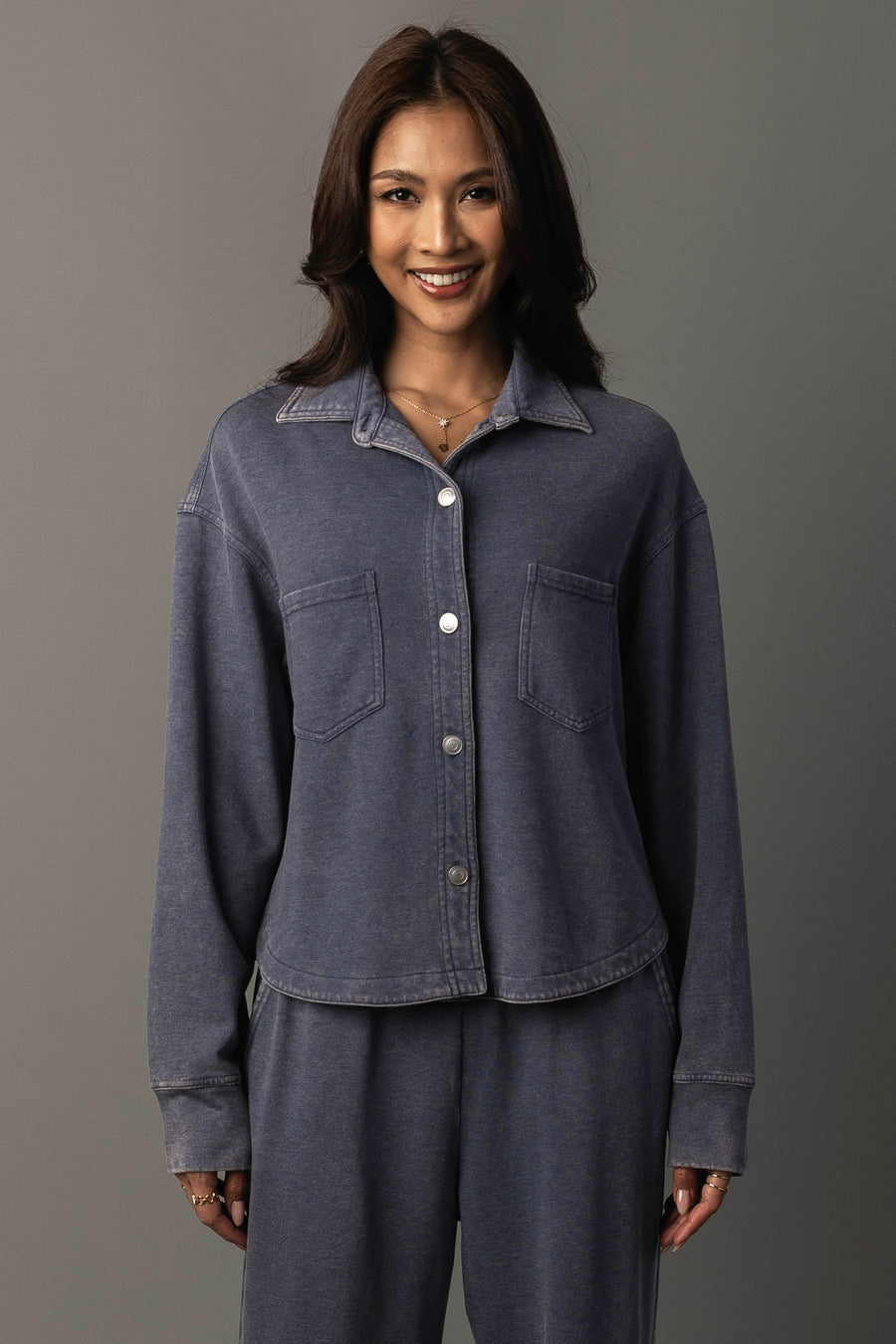 A woman with medium-length dark hair smiles while wearing the Panache Apparel Collection’s Slate Blue Cropped Washed Soft Button Up, paired with matching pants. She stands against a plain gray background.