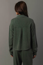 A person with long brown hair, wearing the Panache Apparel Collection Pine Green Cropped Washed Soft Button Up and matching pants, stands facing away from the camera against a plain gray background.