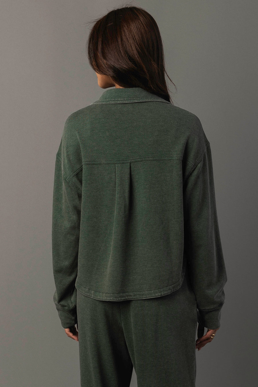 A person with long brown hair, wearing the Panache Apparel Collection Pine Green Cropped Washed Soft Button Up and matching pants, stands facing away from the camera against a plain gray background.