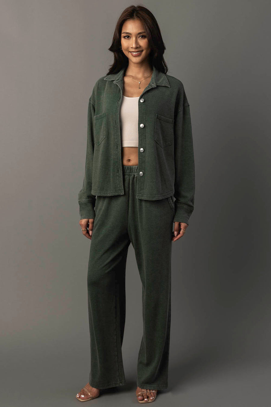 A woman stands smiling against a plain gray background, wearing the Panache Apparel Collection’s Pine Green Cropped Washed Soft Button Up and matching loose pants, layered over a light beige crop top and paired with sandals.