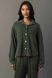A woman with long dark hair and a slight smile stands against a neutral background, wearing the Panache Apparel Collection’s Pine Green Cropped Washed Soft Button Up, styled with matching pants.