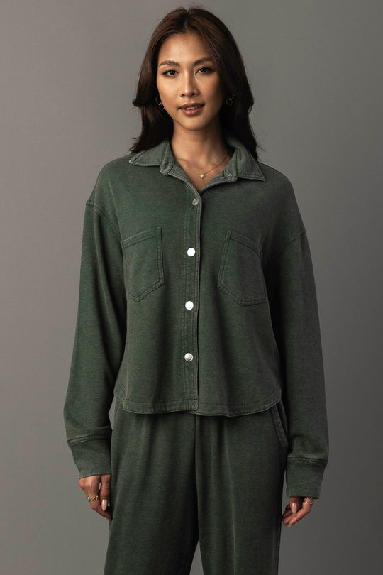 A woman with long dark hair and a slight smile stands against a neutral background, wearing the Panache Apparel Collection’s Pine Green Cropped Washed Soft Button Up, styled with matching pants.