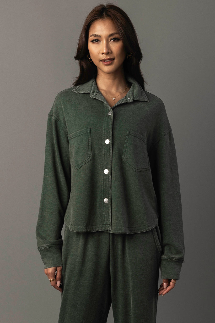 A woman with long dark hair and a slight smile stands against a neutral background, wearing the Panache Apparel Collection’s Pine Green Cropped Washed Soft Button Up, styled with matching pants.