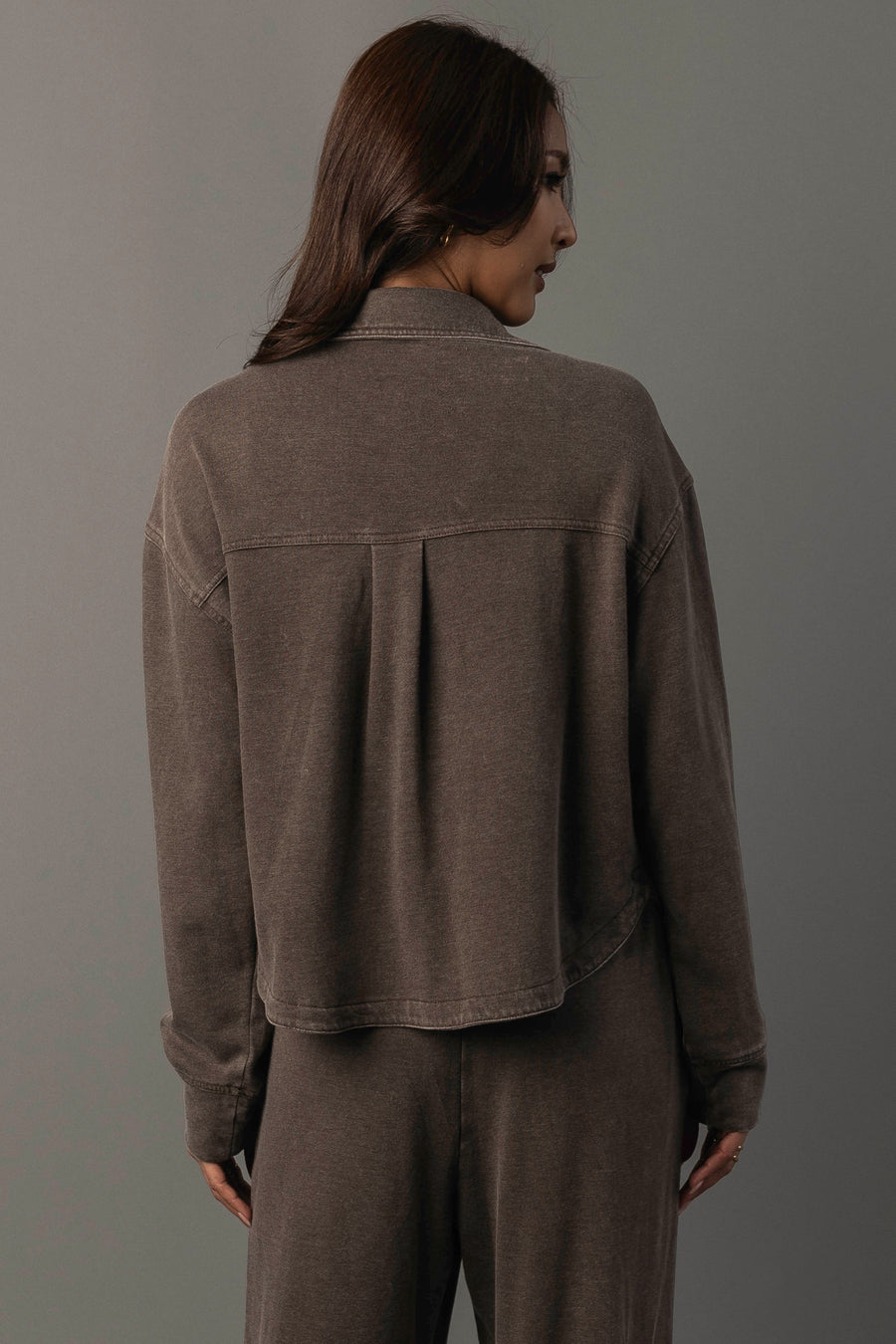 A woman with long brown hair stands facing away against a plain gray background, showcasing the back of the Cocoa Cropped Washed Soft Button Up and matching pants from Panache Apparel Collection.
