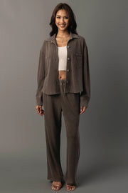 A woman stands smiling against a plain gray background, wearing the Panache Apparel Collection’s Cocoa Cropped Washed Soft Button Up with matching brown pants, a white cropped top, and open-toed heels.