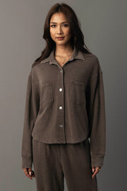 A woman with long brown hair wears the Panache Apparel Collection’s Cocoa Cropped Washed Soft Button Up shirt with matching pants, standing against a plain gray background.