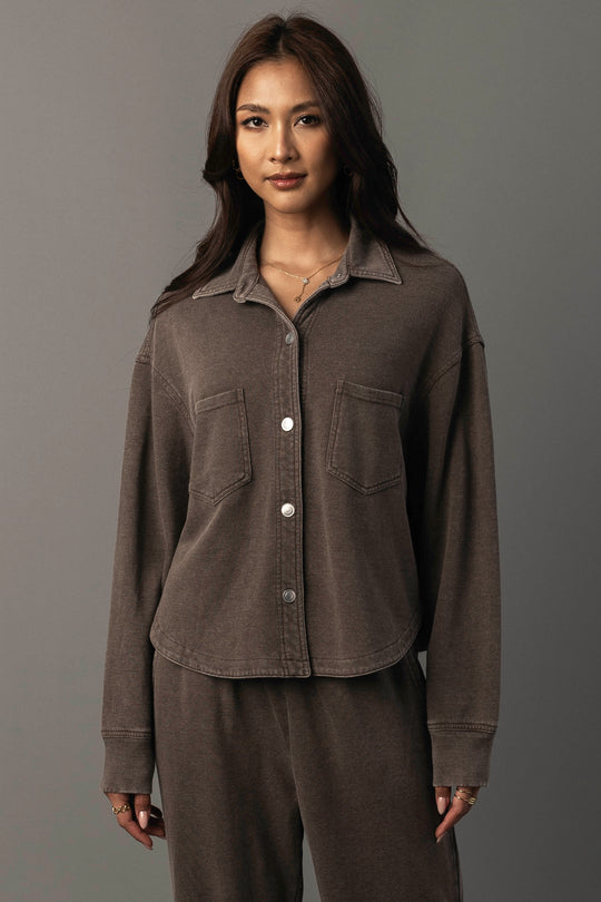 A woman with long brown hair wears the Panache Apparel Collection’s Cocoa Cropped Washed Soft Button Up shirt with matching pants, standing against a plain gray background.