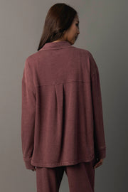 A woman with long brown hair stands facing away, wearing the Merlot Washed Soft Button Up and matching pants from Panache Apparel Collection against a gray background.