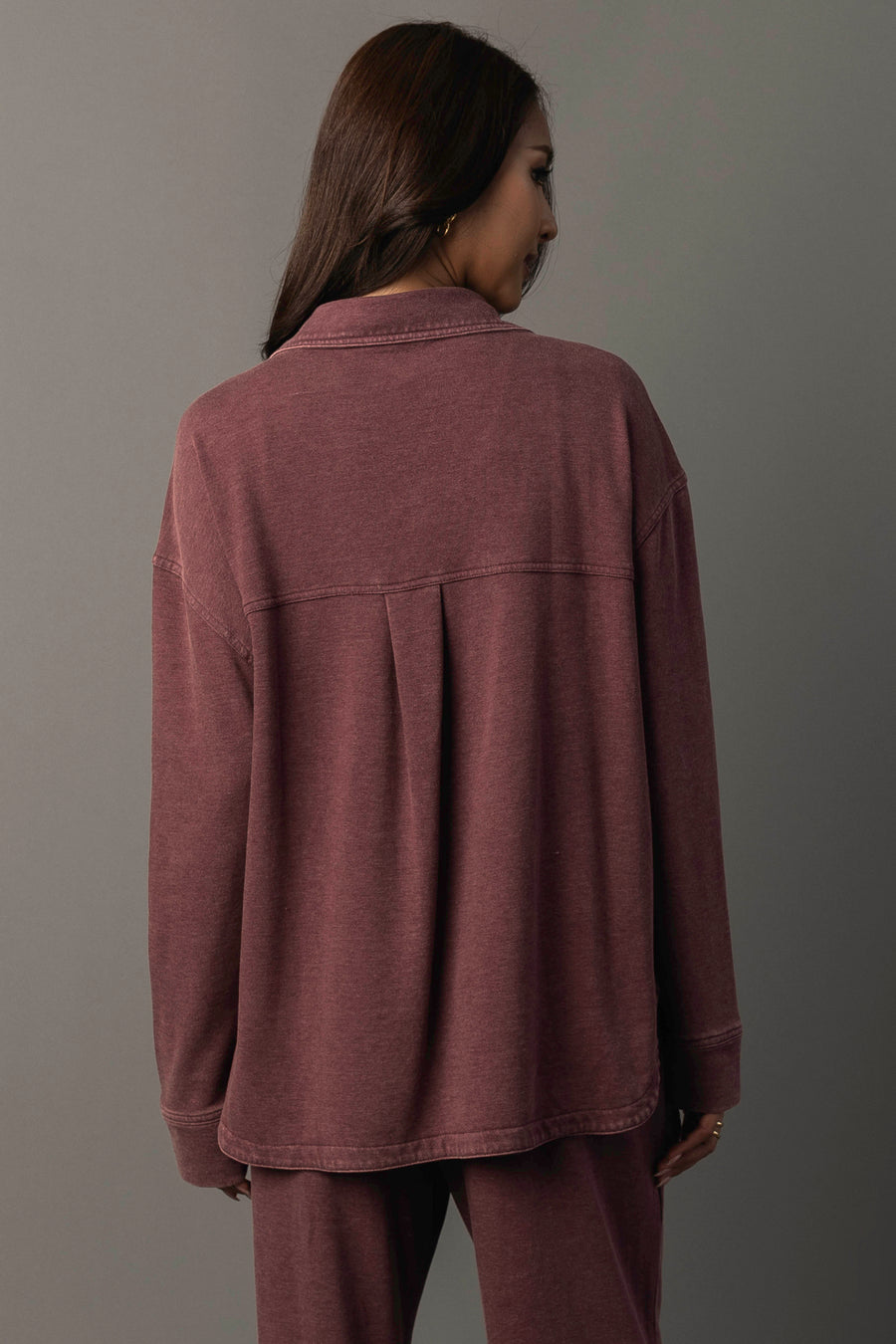 A woman with long brown hair stands facing away, wearing the Merlot Washed Soft Button Up and matching pants from Panache Apparel Collection against a gray background.