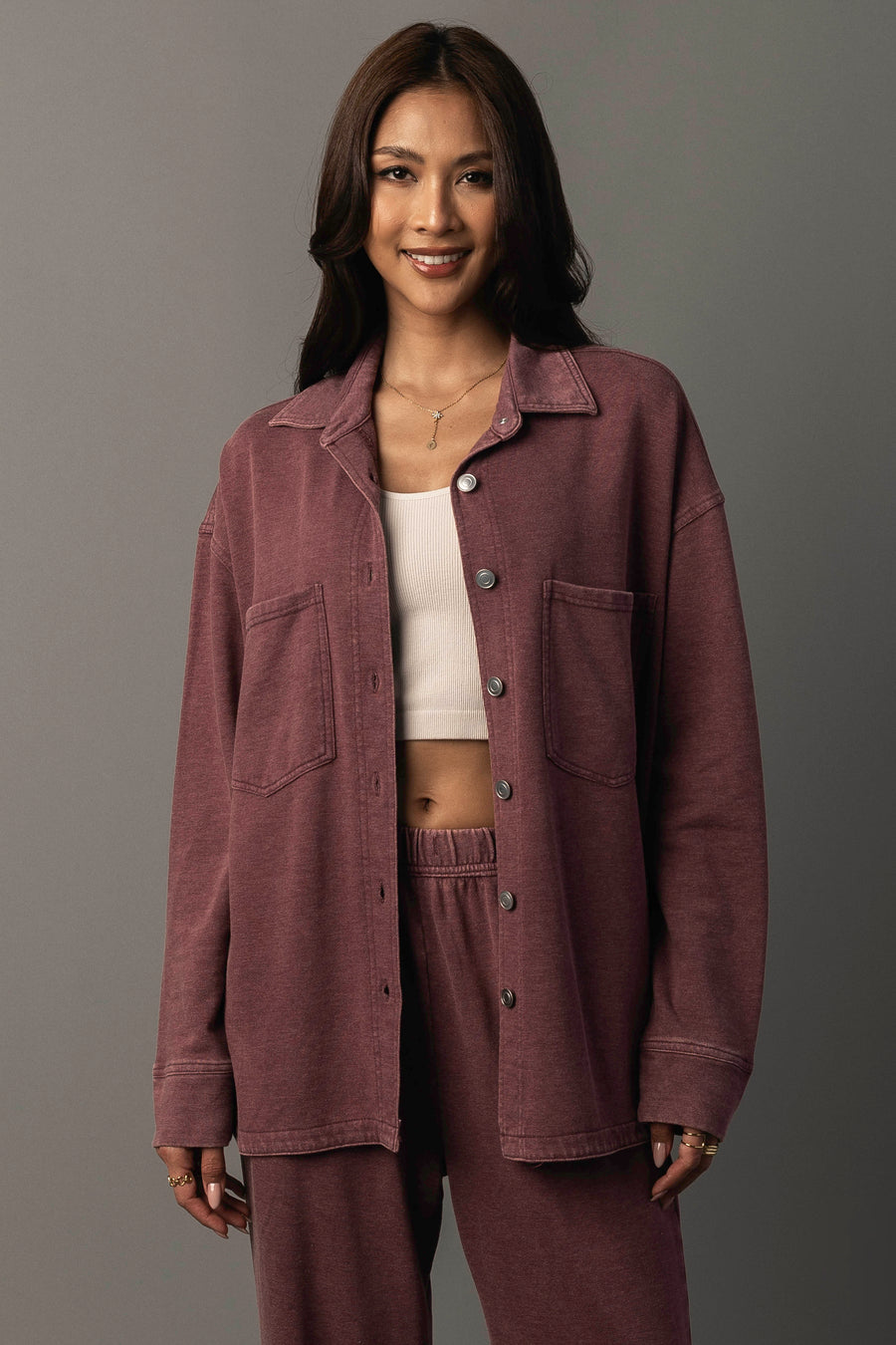 A woman with long brown hair smiles, wearing the Merlot Washed Soft Button Up by Panache Apparel Collection paired with matching pants over a white crop top, standing against a plain gray background.