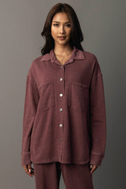A woman wears the Panache Apparel Collection Merlot Washed Soft Button Up, a loose maroon shirt with two chest pockets, paired with matching pants. She stands against a plain gray background, looking at the camera and smiling softly.