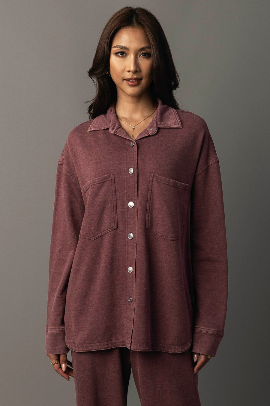 A woman wears the Panache Apparel Collection Merlot Washed Soft Button Up, a loose maroon shirt with two chest pockets, paired with matching pants. She stands against a plain gray background, looking at the camera and smiling softly.
