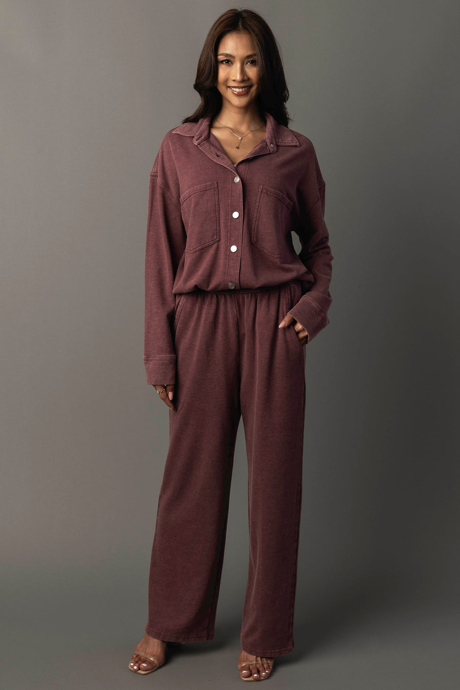 A woman stands smiling with her left hand in her pocket, wearing the Panache Apparel Collection Merlot Washed Soft Button Up and matching wide-leg pants against a plain grey background.