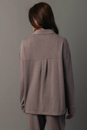 A person with long brown hair stands facing away against a plain gray background, wearing the Panache Apparel Collection Gray Washed Soft Button Up with matching pants.