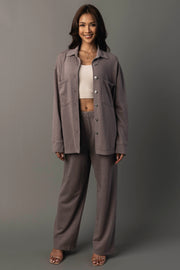 A woman stands smiling against a plain gray background, wearing the Panache Apparel Collection’s Gray Washed Soft Button Up with matching pants layered over a white crop top and finished with open-toe heels.