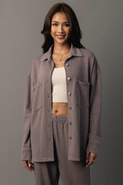 A woman stands smiling, wearing the Panache Apparel Collection Gray Washed Soft Button Up over a white crop top and matching purple pants, against a plain gray background.
